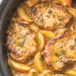 Browned pork chops with apple wedges, sliced onions, and brown sugar sauce in a black slow cooker on a white marble countertop.