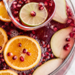 Glass ladle serving sparkling cranberry holiday punch with orange slices, lime, pear, and pomegranate seeds.
