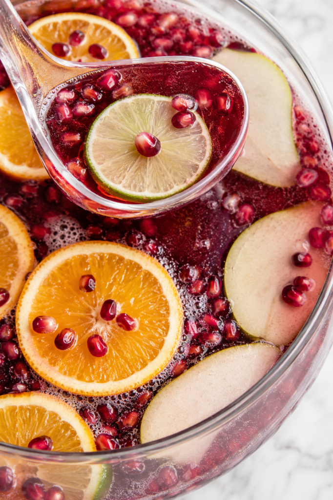 Glass ladle serving sparkling cranberry holiday punch with orange slices, lime, pear, and pomegranate seeds.