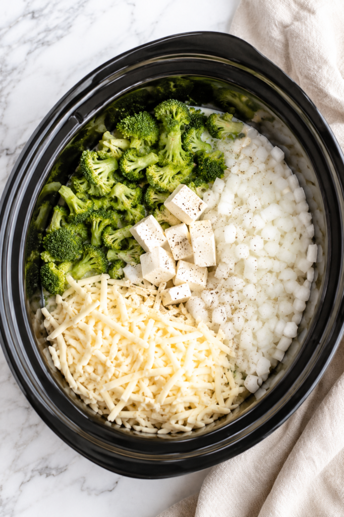 Ingredients for Better Than Panera Crockpot Broccoli Cheddar Soup layered in a slow cooker with fresh broccoli, diced onions, shredded white cheddar, cream cheese, and seasonings.