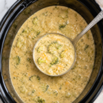 A silver ladle scoops creamy Better Than Panera Crockpot Broccoli Cheddar Soup with tender broccoli from a black slow cooker on a white marble countertop.