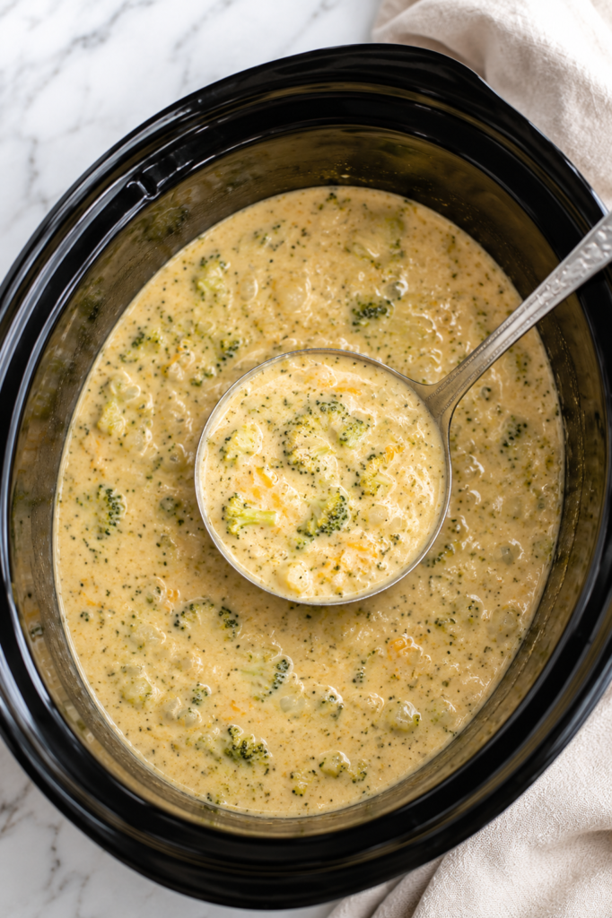 A silver ladle scoops creamy Better Than Panera Crockpot Broccoli Cheddar Soup with tender broccoli from a black slow cooker on a white marble countertop.