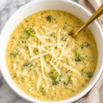 Creamy Better Than Panera Crockpot Broccoli Cheddar Soup in a white bowl topped with shredded white cheddar cheese and served with a gold spoon on a marble countertop.