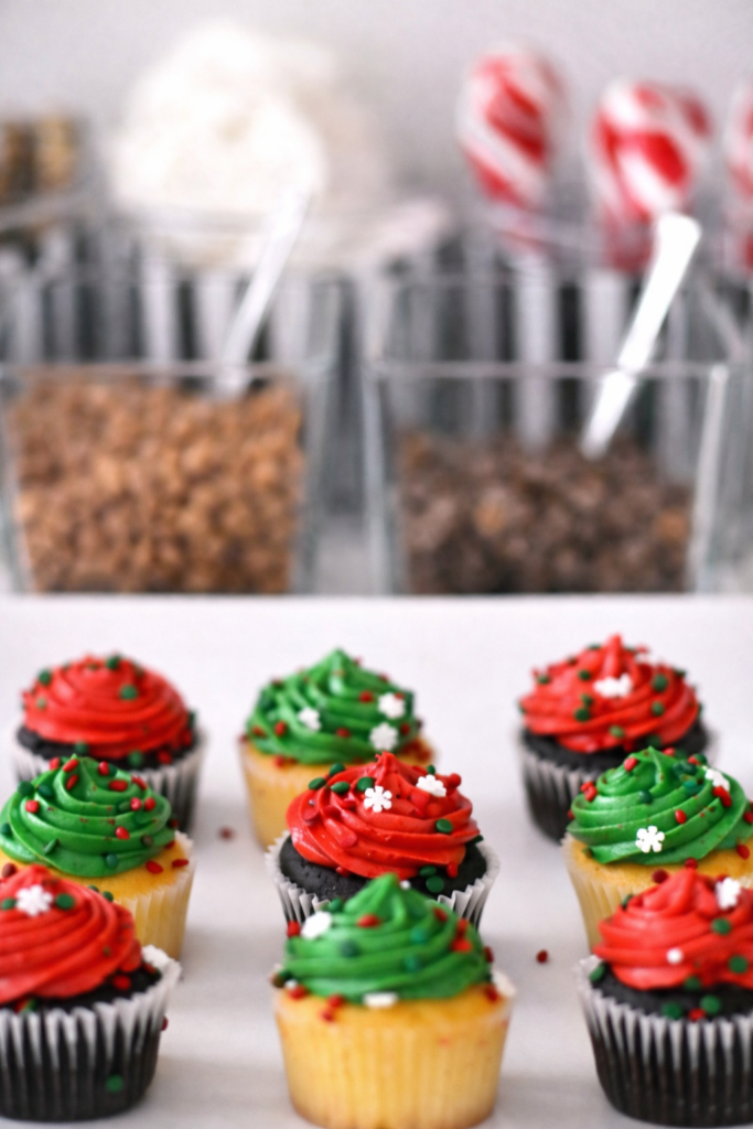 Festive Christmas cupcakes with red and green frosting displayed in front of a holiday hot chocolate bar.