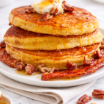 Light and fluffy cornmeal bacon pancakes stacked on a white plate with melted butter and honey dripping down, styled on a marble countertop.