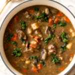 Easy pot roast stew with tender beef, potatoes, carrots and mushrooms in a white Dutch oven with a gold ladle.