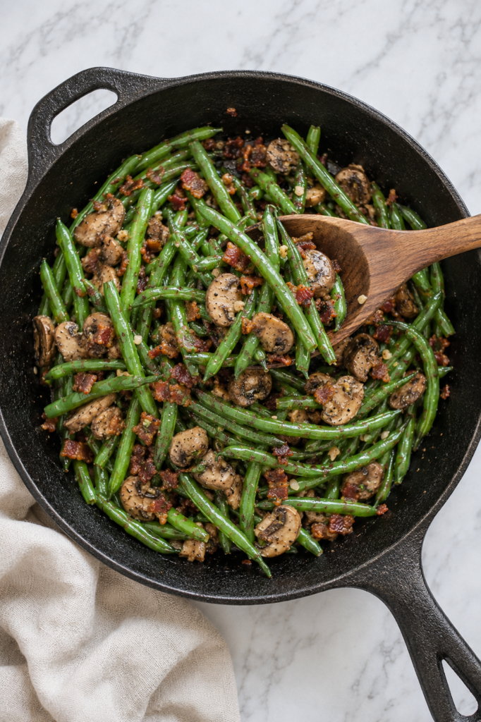 Green beans with bacon and mushrooms being tossed with a wooden spoon in a cast-iron skillet.