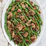 Green beans with bacon and mushrooms served on a white platter with garlic, butter, and cracked black pepper.