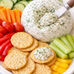 Homemade Boursin cheese ball on a white platter with a cheese spreader scooping cheese onto a cracker, surrounded by cucumbers, tomatoes, carrots, celery, and bell peppers