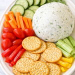 Homemade Boursin cheese ball on a white platter with crackers, cucumber slices, tomatoes, carrots, celery, and bell peppers on a marble counter