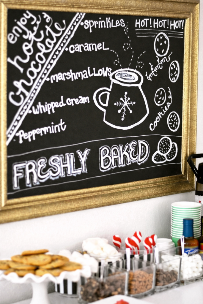 Holiday hot chocolate bar chalkboard sign listing cocoa toppings with jars of marshmallows and cookies below.