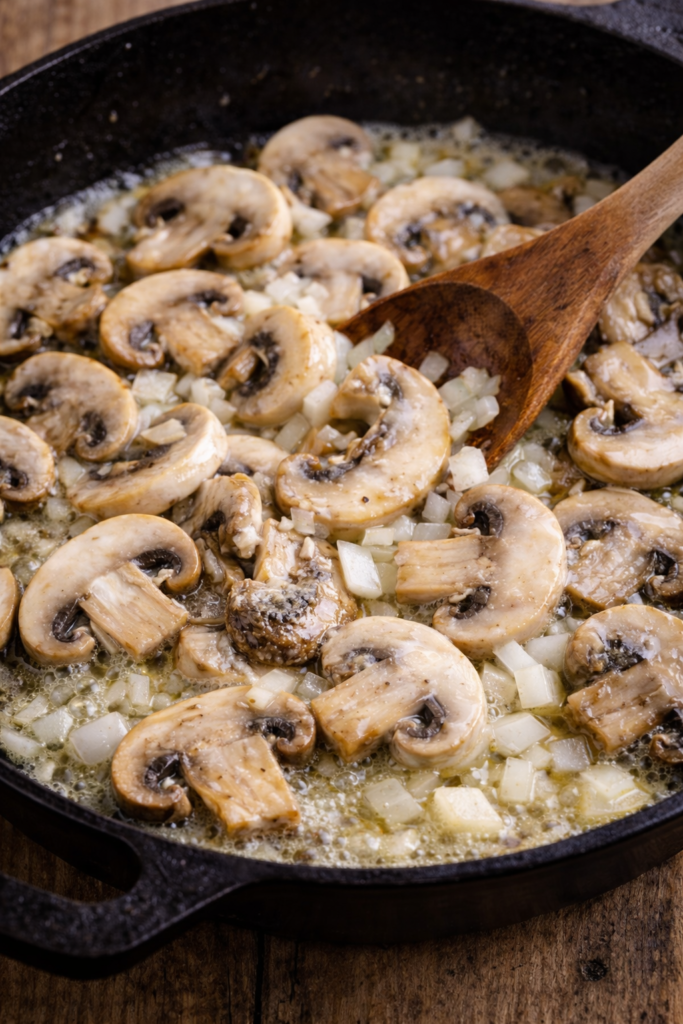Sliced mushrooms and diced onions sautéing in butter in a cast iron skillet with a wooden spoon
