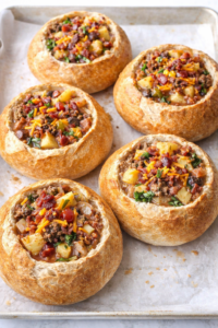 Bacon cheeseburger soup served in homemade bread bowls on a baking sheet, topped with cheddar cheese and crispy bacon