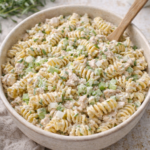 Creamy tarragon tuna pasta salad in a neutral ceramic bowl topped with fresh tarragon and a wooden spoon.