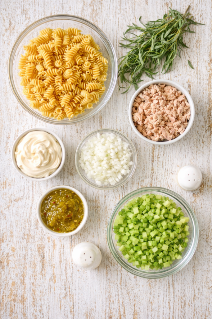 Flat lay of ingredients for tarragon tuna pasta salad including rotini pasta, tuna, celery, onion, mayonnaise, relish, and fresh tarragon.