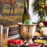 Holiday bubbly bar with Prosecco in an antique silver ice bucket, bowls of fruit, equestrian artwork, and lush lighted Christmas garland.