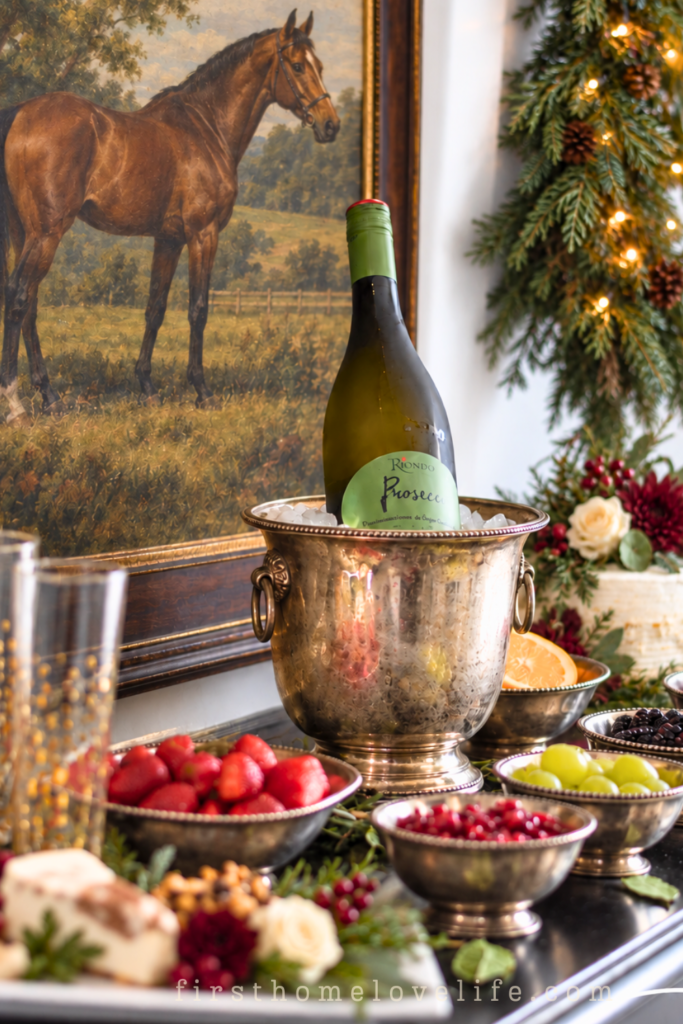 Holiday bubbly bar with Prosecco in an antique silver ice bucket, bowls of fruit, equestrian artwork, and lush lighted Christmas garland.