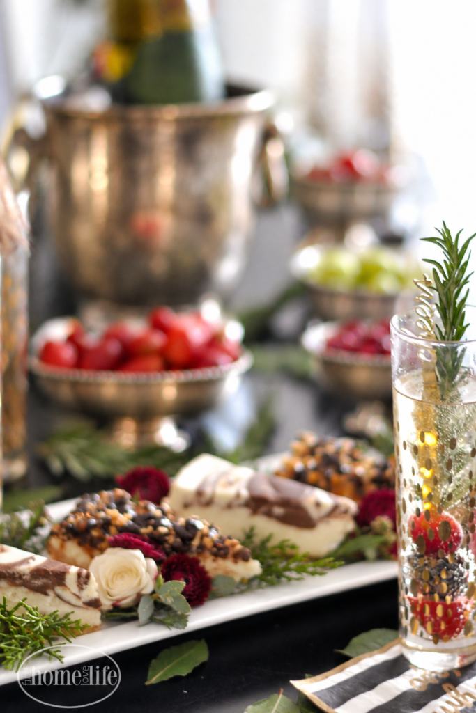 Sparkling wine with berries and rosemary beside a dessert platter with ivory and burgundy flowers, with antique silver fruit bowls behind.
