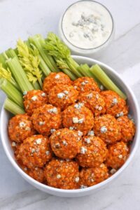 Baked buffalo chicken ball bites topped with blue cheese and celery in white bowl