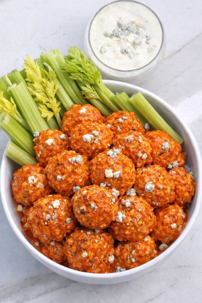 Baked buffalo chicken ball bites topped with blue cheese and celery in white bowl