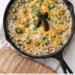Hot roast beef dip baked in a cast iron skillet with melted cheddar cheese and pickles, served with sliced rye bread on a neutral linen background.