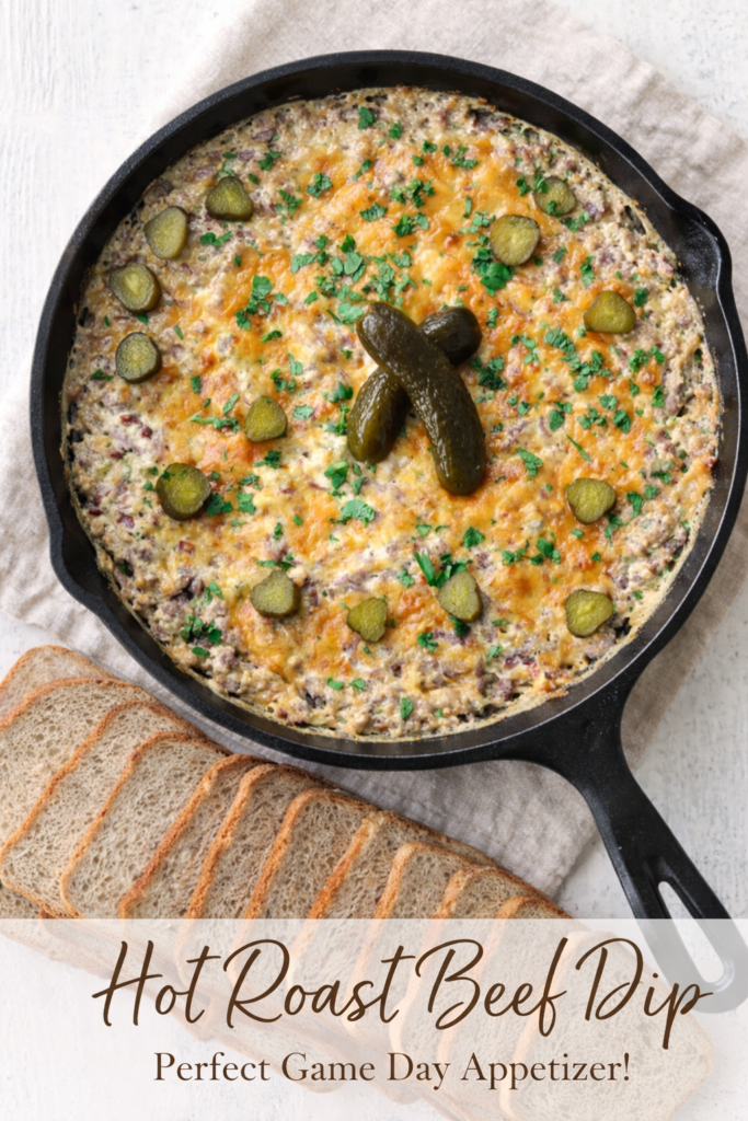 Hot roast beef dip baked in a cast iron skillet with melted cheddar cheese and pickles, served with sliced rye bread on a neutral linen background.