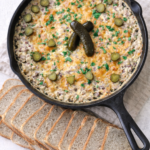 Hot roast beef dip baked in a cast iron skillet with melted cheddar and pickles, served with sliced rye bread on a light neutral background.