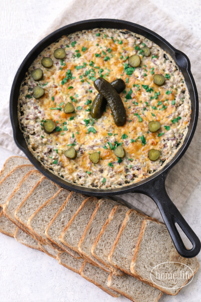 Hot roast beef dip baked in a cast iron skillet with melted cheddar and pickles, served with sliced rye bread on a light neutral background.