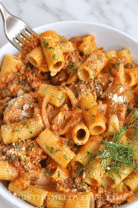 Fork lifting a bite of creamy Italian sausage rigatoni with fennel from a white bowl.