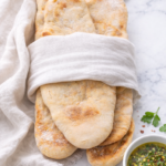Soft homemade pita bread wrapped in linen on a marble countertop with a bowl of herbed olive oil for dipping.