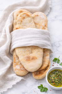 Soft homemade pita bread wrapped in linen on a marble countertop with a bowl of herbed olive oil for dipping.