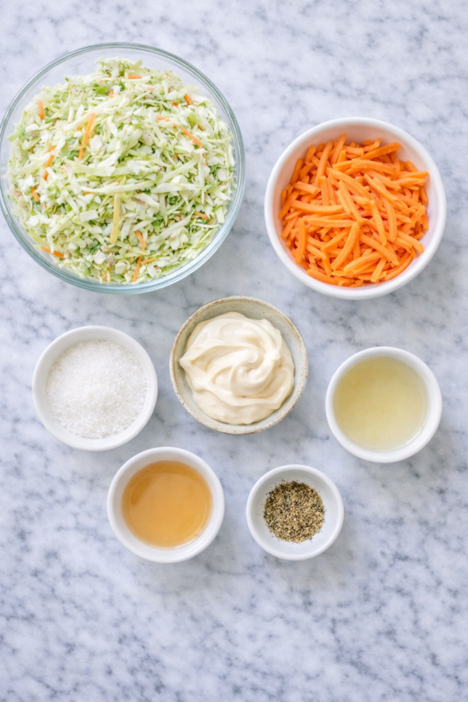 Flat lay of creamy sweet coleslaw ingredients including shredded cabbage, carrots, mayonnaise, sugar, vinegar, and seasonings on a white marble countertop