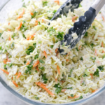 Creamy sweet coleslaw being tossed with tongs in a glass bowl with cabbage, carrots, and homemade dressing