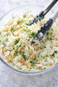 Creamy sweet coleslaw being tossed with tongs in a glass bowl with cabbage, carrots, and homemade dressing