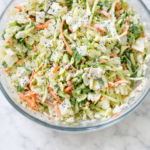 Creamy sweet coleslaw in a glass bowl with shredded cabbage, carrots, and poppy seed dressing on a white marble counter