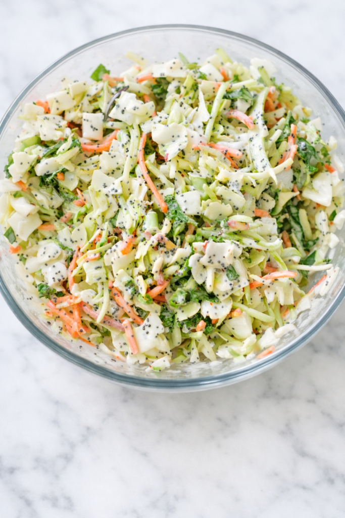 Creamy sweet coleslaw in a glass bowl with shredded cabbage, carrots, and poppy seed dressing on a white marble counter