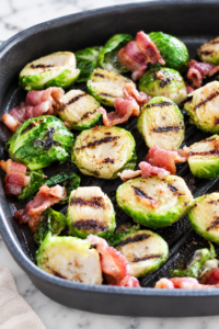 Grilled Brussels sprouts with browned edges and bacon pieces in a black grill pan on a white marble countertop.