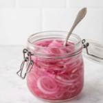 Quick pickled red onions in a glass jar on a white marble countertop with a white tile background.