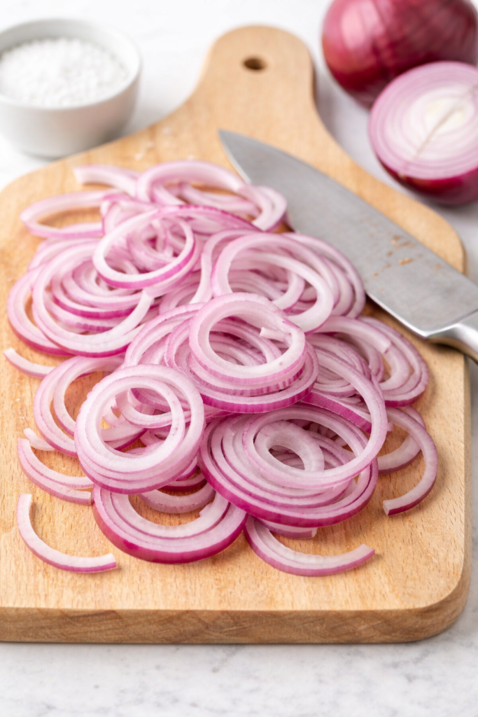 Thinly sliced red onions on a wooden cutting board with a chef’s knife.
