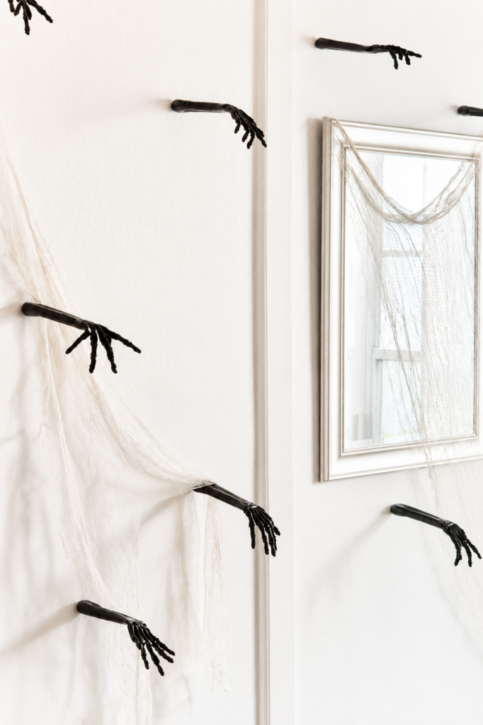 Black skeleton hands emerging from a white wall with draped gauze and a vintage mirror creating elegant Halloween wall decor.