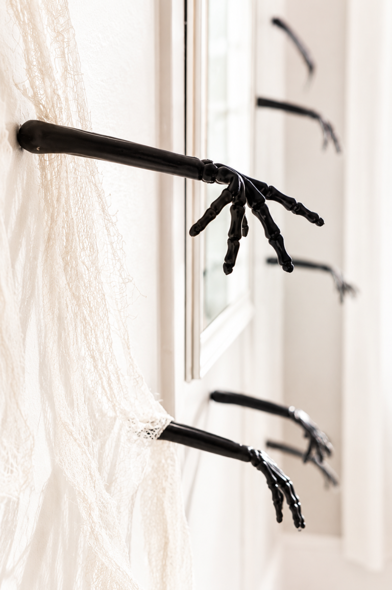 Close-up of black skeleton hands emerging from a white wall with draped gauze fabric for elegant Halloween decor.