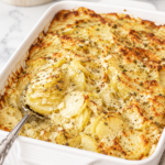 Creamy cheesy scalloped potatoes baked until golden brown in a white rectangular baking dish with a silver serving spoon.