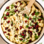Creamy white cheddar macaroni and cheese topped with bright dried cranberries and fresh herbs in a white enamel casserole dish on a marble counter.