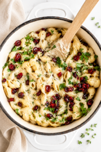 Creamy white cheddar macaroni and cheese topped with bright dried cranberries and fresh herbs in a white enamel casserole dish on a marble counter.