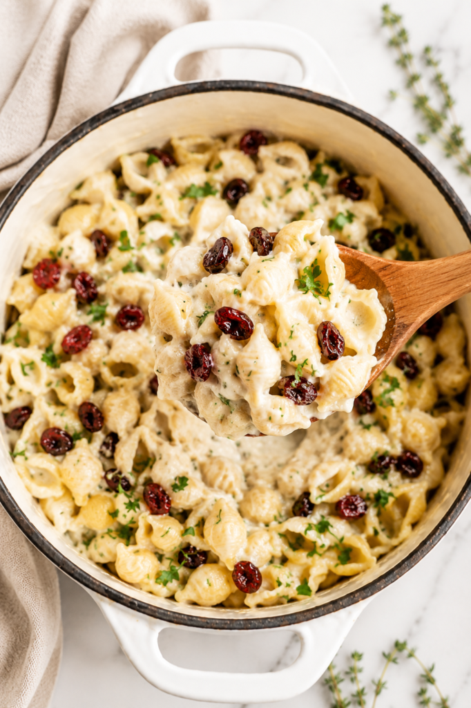 Wooden spoon lifting a creamy scoop of white cheddar cranberry mac and cheese from a white enamel casserole dish with dried cranberries and fresh herbs.