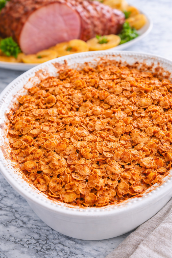cheesy cornflake potato casserole in white baking dish on marble counter with crispy topping and blurred baked ham in background