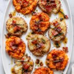 Crispy smashed sweet potatoes topped with bacon, fresh rosemary, sage, and a buttery maple glaze on a white platter over a marble countertop.