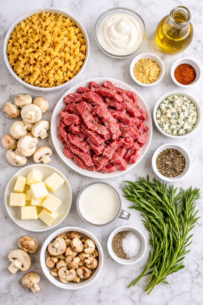 Flat lay of ingredients for blue cheese beef stroganoff including steak, mushrooms, egg noodles, sour cream, and blue cheese on a marble counter