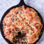 Breakfast sausage, bacon, and egg biscuit casserole baked in a cast iron skillet on a white marble countertop
