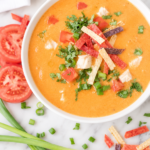 Creamy Chili's copycat chicken enchilada soup served in a white bowl on a marble countertop with tortilla strips, tomatoes, cilantro, and green onions.